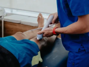 Close-up of a healthcare provider in blue scrubs applying a handheld ultrasound or shockwave therapy device to the back of a patient's leg. The patient is lying face down on a treatment table wearing blue athletic shorts.