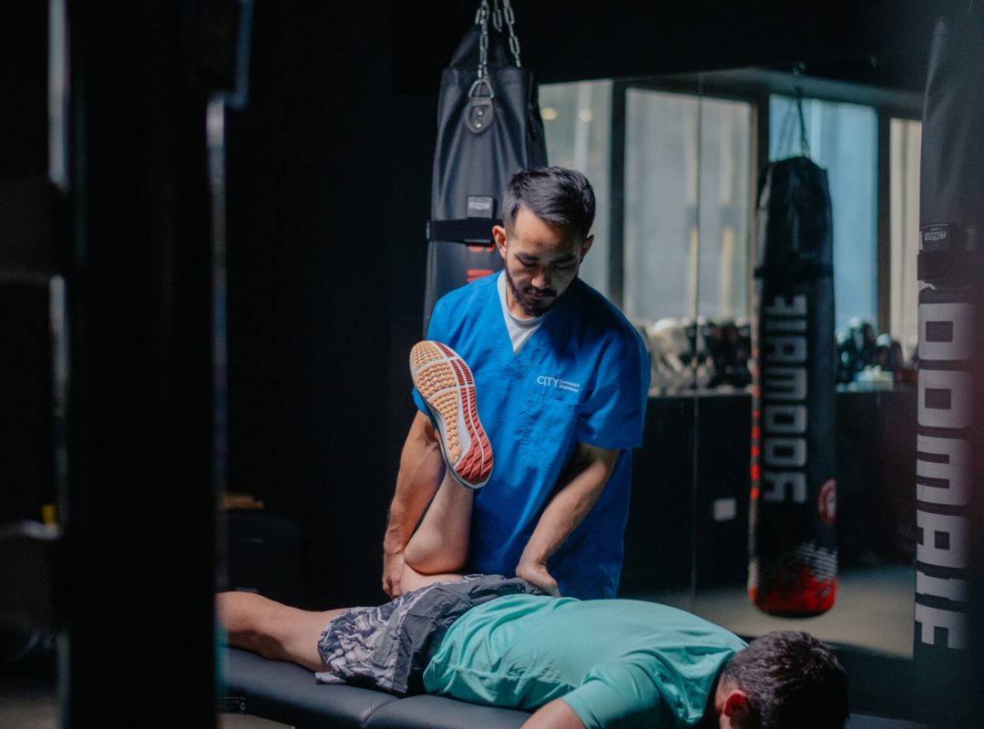 An osteopath in a blue shirt examining a runner lying on a therapy table in a dimly lit room with fitness equipment in the background.